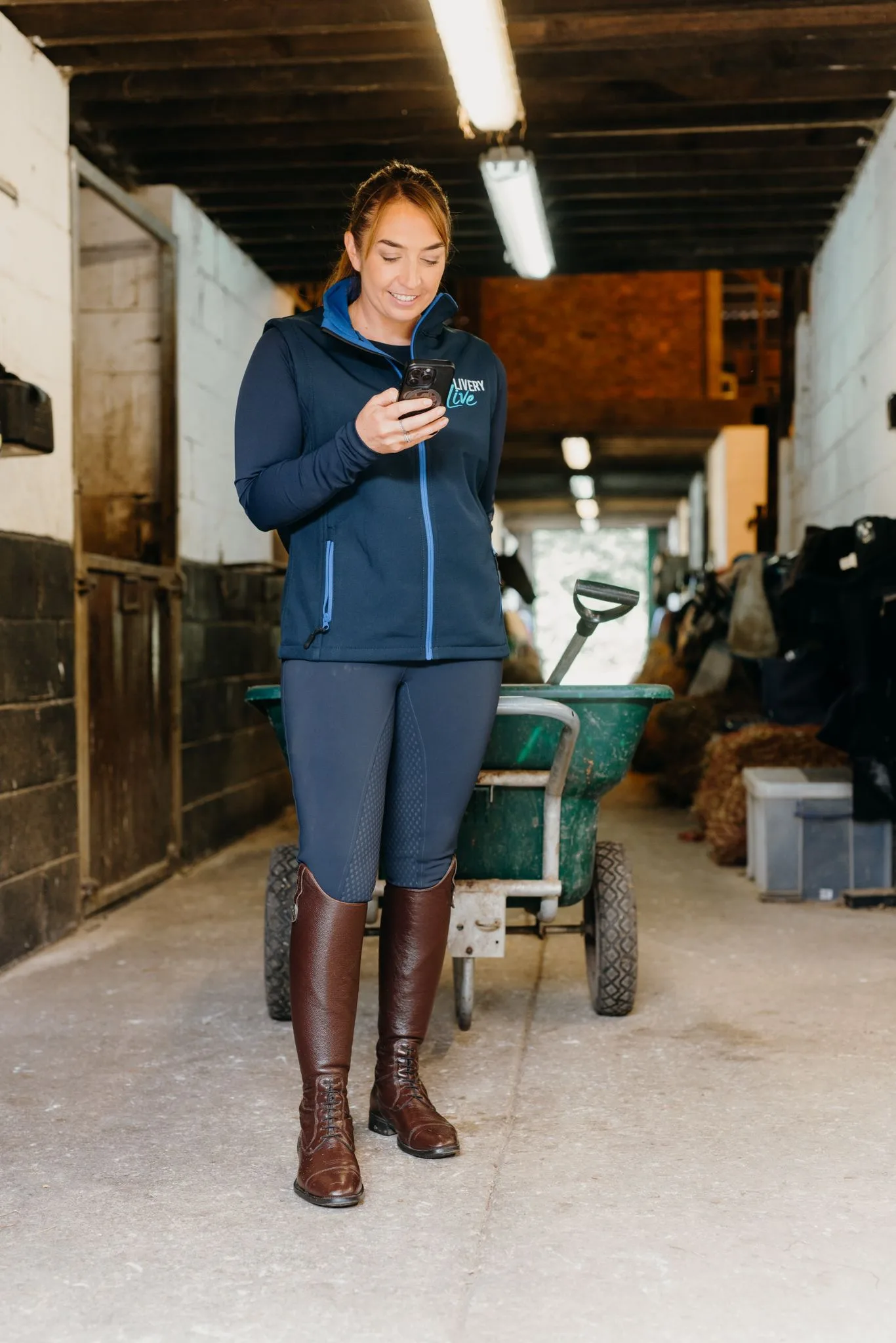 Image shows a lady in horses stables looking at her phone. Image depicts how LIVERYLive helps you go from yard work to invoices