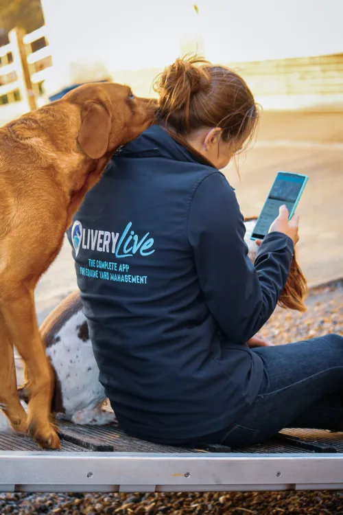 Image shows a lady sitting looking at the LIVERYLive app on her phone with a dog resting it's head on her shoulder
