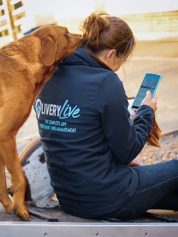 Image shows a lady sitting looking at the LIVERYLive app on her phone with a dog resting its head on her shoulders. Image depicts Arena Booking Software for Livery Yards, Riding Arenas & Equestrian Centres